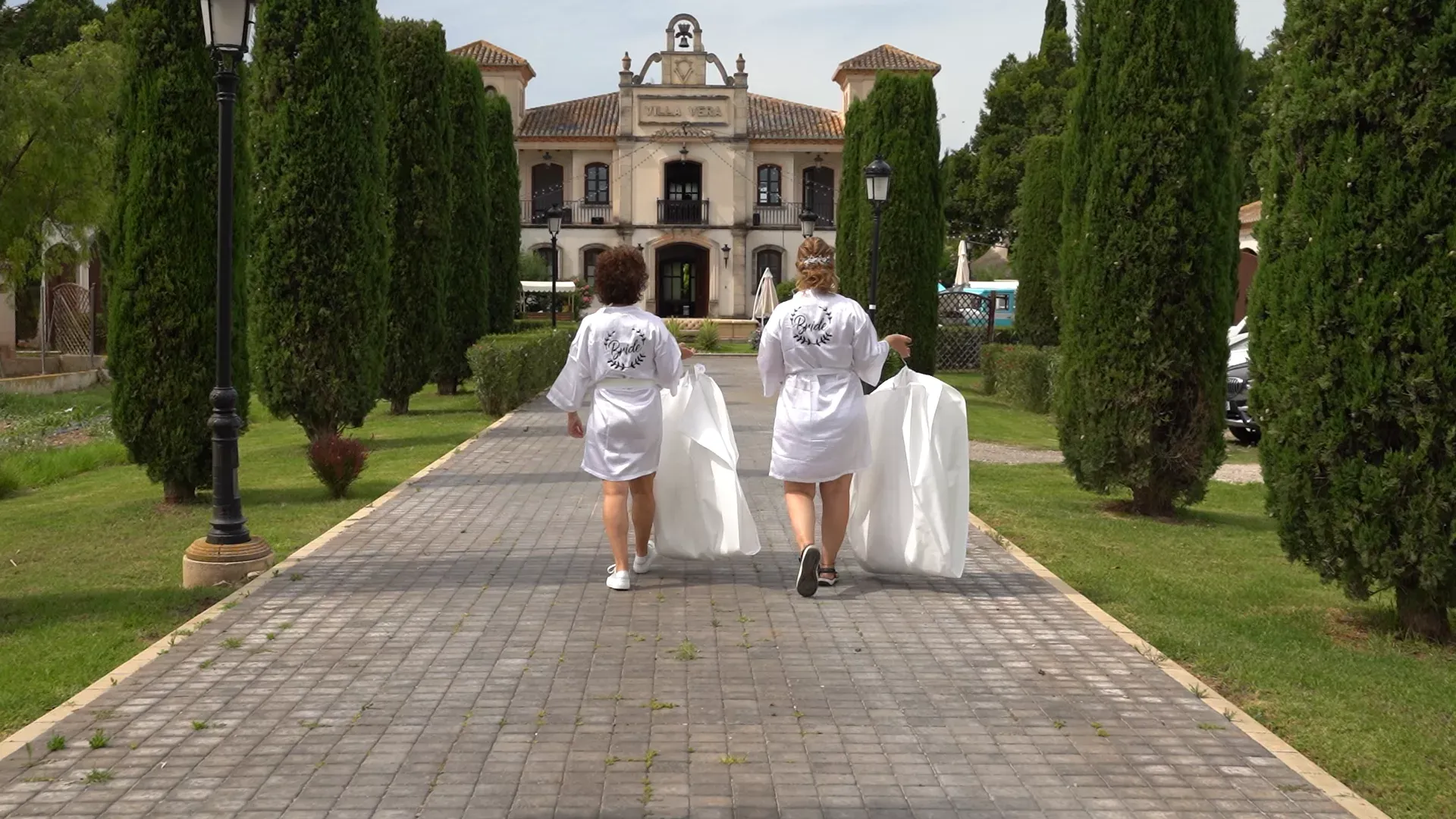 Novia y dama de honor en albornoz subiendo por un camino arbolado hacia una casa señorial