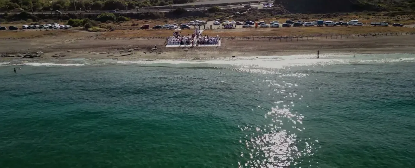 Aerial view over the turquoise Mediterranean towards a beach wedding ceremony