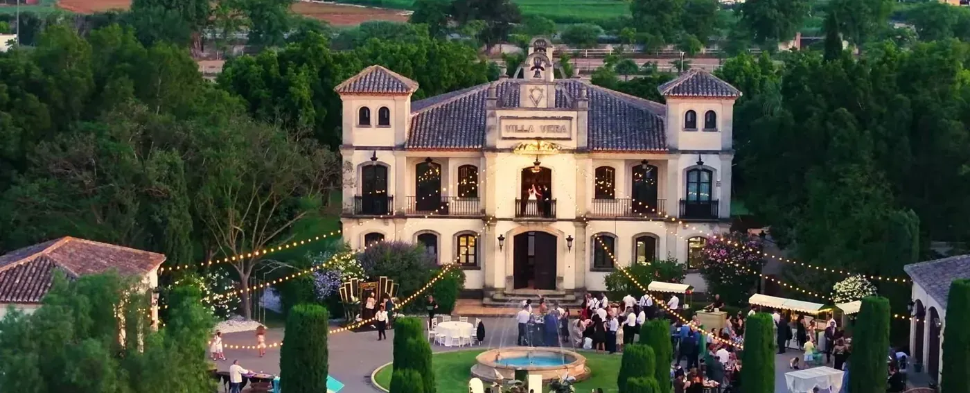 Aerial view of a palace-style finca at dusk with lights glowing across the garden